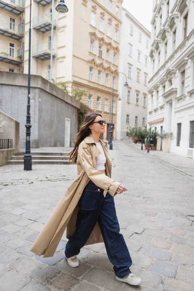 Longitud completa de la mujer joven en gafas de sol de moda y abrigo de trinchera caminando en la calle de Viena - foto de stock