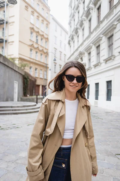 Mujer joven y complacida en gafas de sol de moda y abrigo de trinchera caminando por la calle de Viena - foto de stock