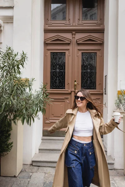 Cheerful young woman in trendy sunglasses and trench coat holding paper cup near entrance door — Stock Photo