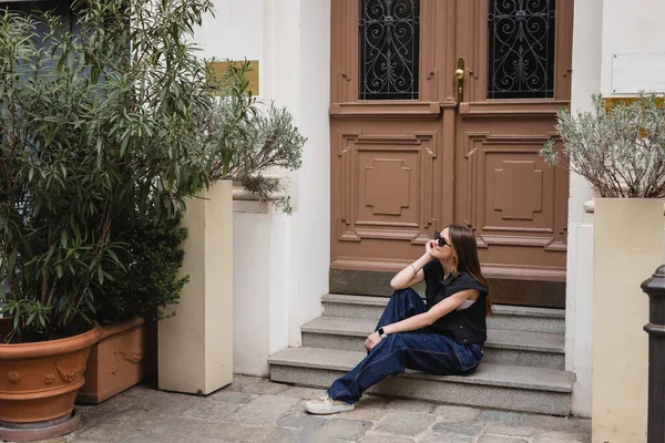 Volle Länge der fröhlichen jungen Frau in trendiger Sonnenbrille und ärmelloser Jacke, die auf der Treppe neben der Eingangstür sitzt — Stockfoto