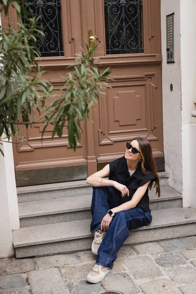 Pleine longueur de jeune femme heureuse en lunettes de soleil et veste sans manches assis sur les escaliers près de la porte d'entrée — Photo de stock