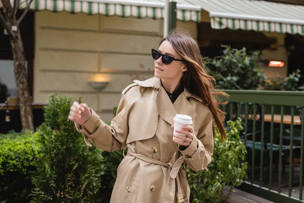 Young woman in trendy sunglasses and trench coat holding paper cup — Stock Photo