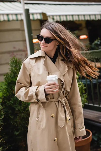 Glückliche junge Frau in stylischer Sonnenbrille und Trenchcoat mit Pappbecher — Stockfoto