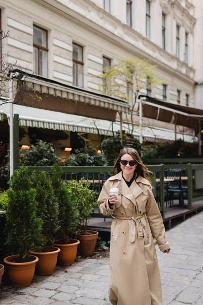 Jeune femme souriante dans des lunettes de soleil élégantes et trench coat tenant tasse en papier à l'extérieur — Photo de stock