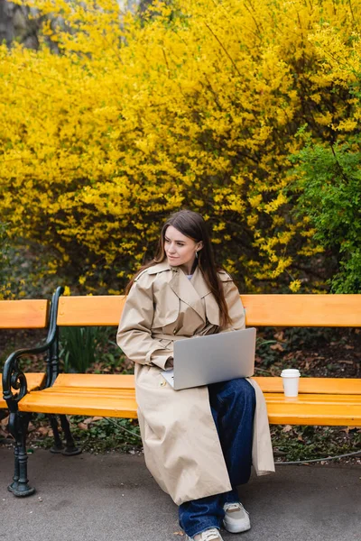 Young woman in beige trench coat sitting on bench with laptop near paper cup — Stock Photo