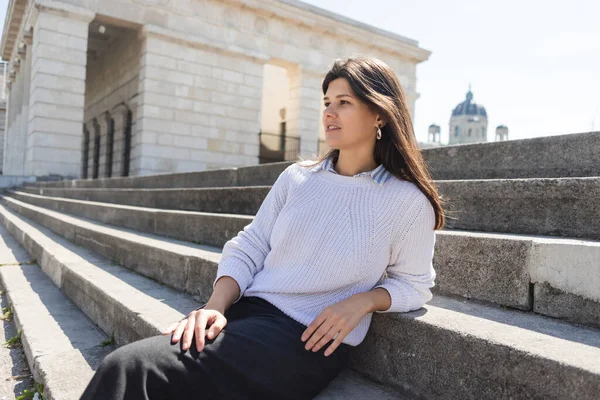 Brunette woman looking away and sitting on concrete stairs in vienna — Stock Photo