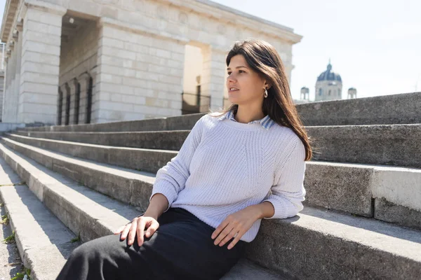 Morena mujer mirando hacia otro lado y sentado en escaleras de hormigón en Viena - foto de stock