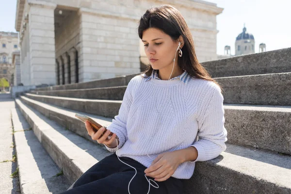 Brunette woman listening music in wired earphones and using smartphone while sitting on stairs — Stock Photo