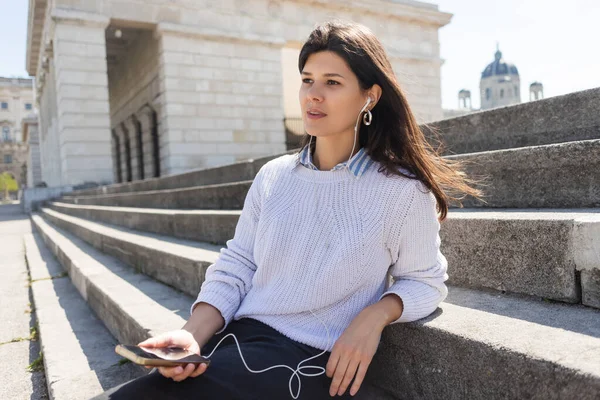 Brunette woman listening music in wired earphones and holding smartphone while sitting on stairs — Stock Photo
