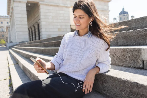 Femme heureuse écouter de la musique dans les écouteurs filaires et en utilisant un smartphone tout en étant assis sur les escaliers — Photo de stock