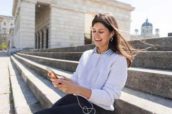Весела жінка слухає музику в дротових навушниках і використовує смартфон, сидячи на сходах — стокове фото