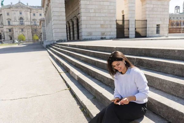 Mujer sonriente escuchando música en auriculares con cable y usando un teléfono inteligente mientras está sentada en las escaleras - foto de stock