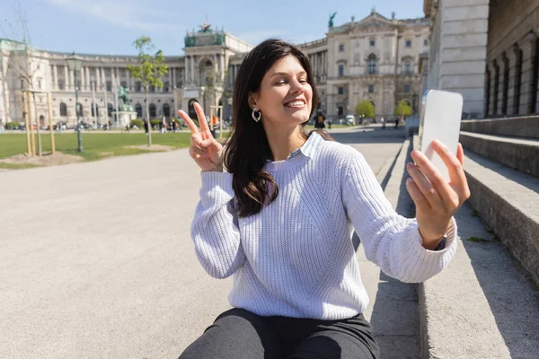 Mujer feliz mostrando señal de paz mientras toma selfie en el teléfono inteligente en Viena - foto de stock