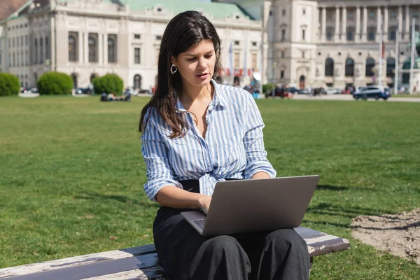 Brunette freelancer за допомогою ноутбука під час сидіння на лавці в зеленому парку vienna — стокове фото