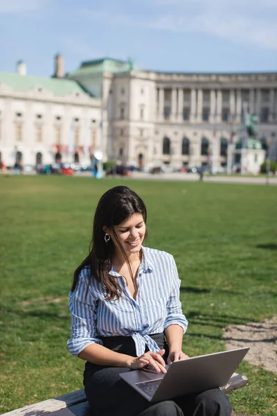 Feliz freelancer usando el ordenador portátil mientras está sentado en el banco en el parque verde de Viena - foto de stock
