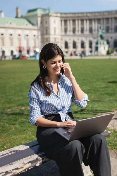 Щасливий фрилансер розмовляє на смартфоні і за допомогою ноутбука, сидячи на лавці в зеленому парку венни. — стокове фото