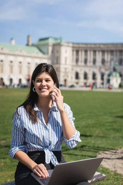 Smiling freelancer talking on smartphone and using laptop while sitting on bench in green park of vienna — Stock Photo