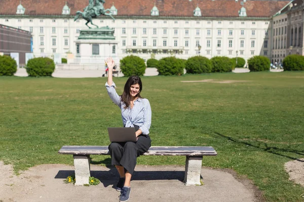 Brünette Frau sitzt mit Laptop auf Bank und winkt mit der Hand im grünen Park von Wien — Stockfoto
