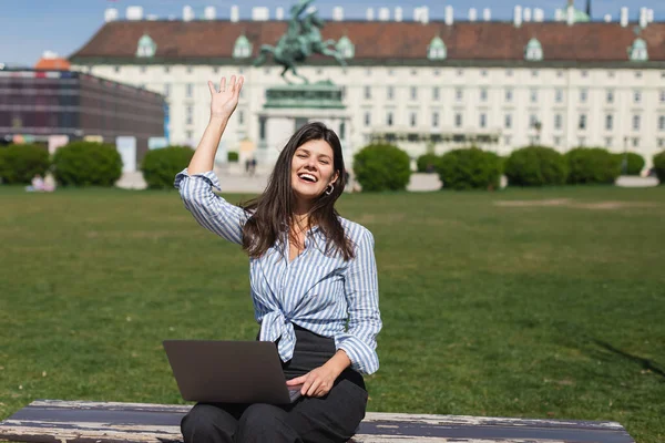 Щаслива жінка сидить на лавці з ноутбуком і махає рукою в зеленому парку венни. — стокове фото