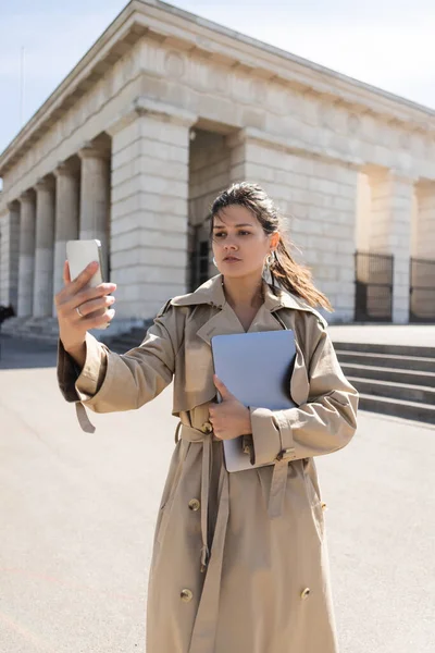 Brunette woman in trench coat holding laptop and looking at smartphone outside — Stock Photo