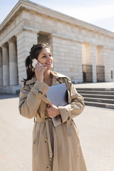 Happy woman in trench coat holding laptop and talking on smartphone outside — Stock Photo