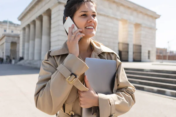 Cheerful woman in trench coat holding laptop and talking on smartphone outside — Stock Photo