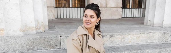 Pleased woman in trench coat smiling and sitting on concrete stairs, banner — Stock Photo