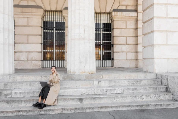 Lebensfrohe Frau im Trenchcoat, die auf Betontreppen sitzt, am Smartphone spricht und in Wien ferngesteuert am Laptop arbeitet — Stockfoto