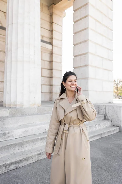 Femme heureuse en trench coat beige parlant sur smartphone dans la rue près du bâtiment de vienne — Photo de stock