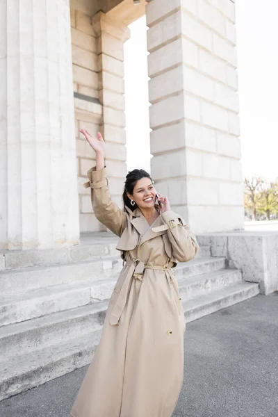 Mujer feliz en gabardina beige agitando la mano mientras habla en el teléfono inteligente en la calle cerca del edificio de Viena - foto de stock