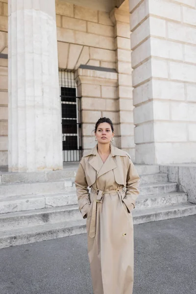 Brünette Frau im beigen Trenchcoat blickt in die Kamera, während sie mit den Händen in den Taschen in der Nähe des Gebäudes von Wien steht — Stockfoto