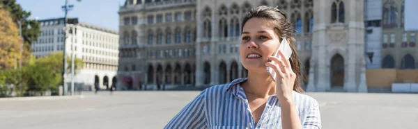 Cheerful woman in striped shirt talking on mobile phone on street in vienna, banner — Stock Photo