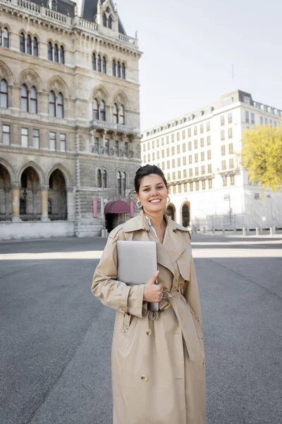 Happy freelancer in beige trench coat walking with laptop on street of vienna — Stock Photo