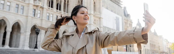 Mujer feliz en gabardina beige tomando selfie cerca del ayuntamiento de Viena, pancarta - foto de stock