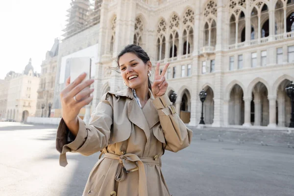 Mulher feliz em casaco de trincheira bege mostrando sinal de paz tomando selfie perto da prefeitura de Viena — Fotografia de Stock