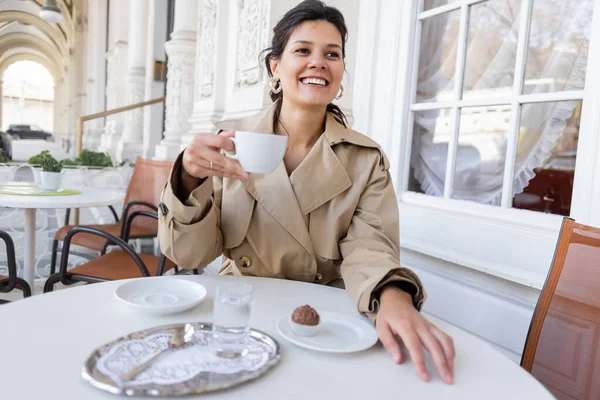 Happy woman in trench coat sitting near cupcake and holding cup of coffee in cafe terrace — Stock Photo