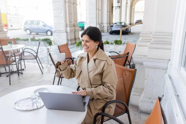 Mujer feliz en gabardina usando el ordenador portátil y sosteniendo la taza mientras está sentado en la terraza de la cafetería en la calle en Viena - foto de stock