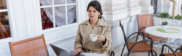 Woman in trench coat using laptop and holding cup in cafe terrace on european street in vienna, banner — Stock Photo