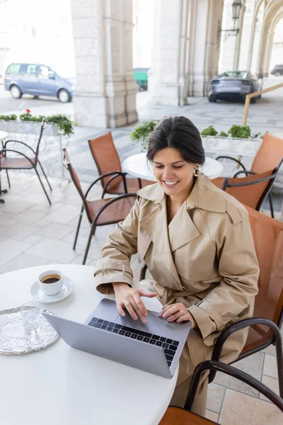 Веселая женщина в плаще с ноутбуком на террасе кафе на европейской улице — стоковое фото