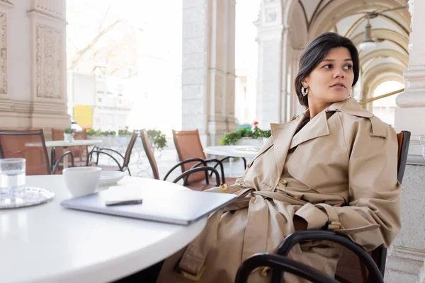 Woman in trench coat sitting near laptop and drinks in cafe terrace on european street in vienna — Stock Photo