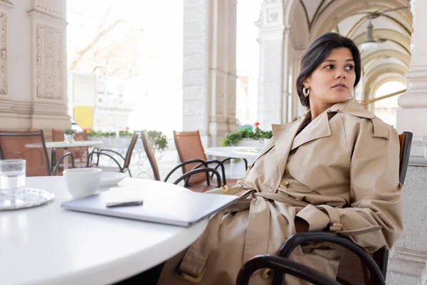 Femme en trench coat assis près d'un ordinateur portable et boissons dans la terrasse du café sur la rue européenne à Vienne — Photo de stock