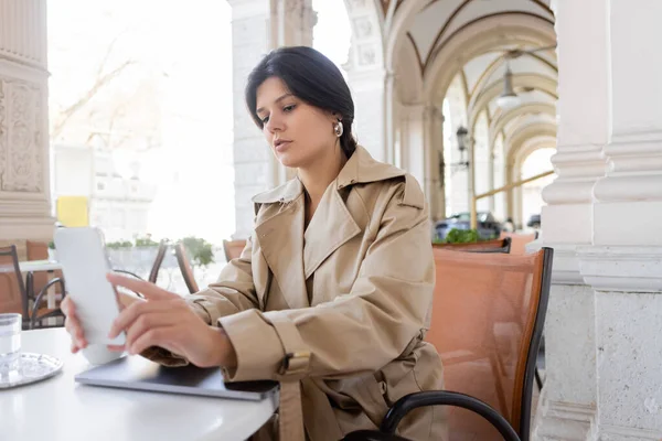 Freelancer en gabardina sosteniendo smartphone cerca de laptop en cafetería terraza en calle europea en vienna - foto de stock