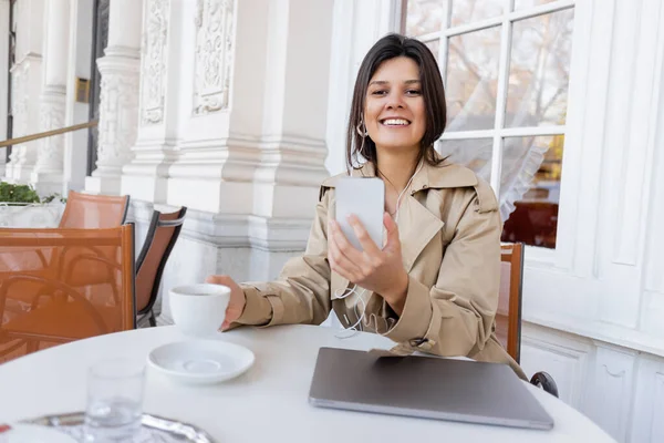 Mujer sonriente en gabardina escuchando música en auriculares y sosteniendo la taza mientras usa el teléfono inteligente en la terraza de la cafetería - foto de stock