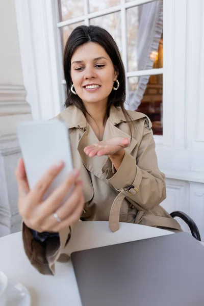 Cheerful woman in trench coat gesturing while taking selfie on blurred smartphone in cafe terrace — Stock Photo