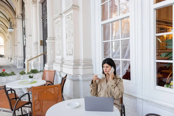 Freelancer in trench coat talking on cellphone near laptop and holding cup of coffee on cafe terrace in vienna — Stock Photo
