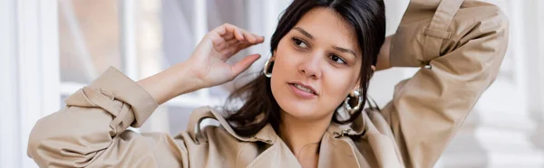 Retrato de mujer en gabardina beige y pendientes de aro ajustando el cabello exterior, bandera - foto de stock