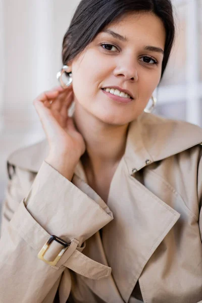 Ritratto di donna felice in trench beige e orecchini a cerchio sorridenti fuori — Foto stock