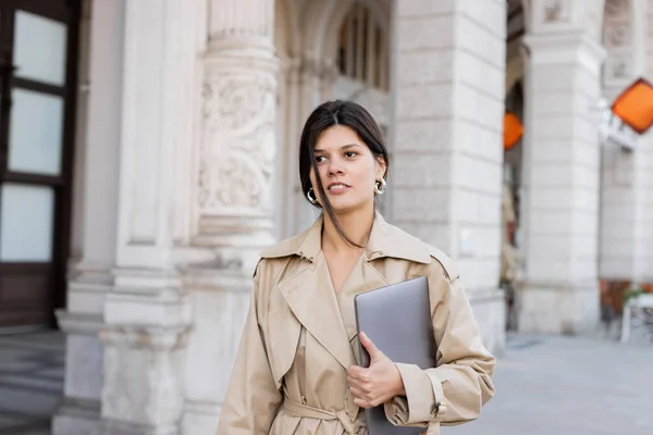 Stylish woman in beige trench coat walking with laptop on street of vienna — Stock Photo