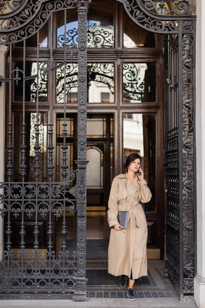 Жінка в траншеї пальто говорить на смартфоні і стоїть з ноутбуком біля кованих дверей — стокове фото