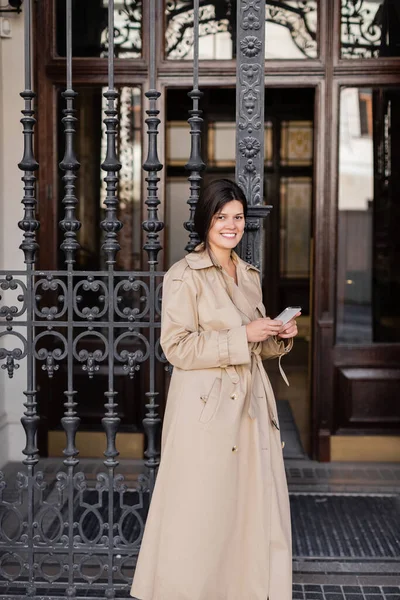 Glückliche Frau im Trenchcoat hält Smartphone in der Hand und steht vor gefälschten Türen — Stockfoto